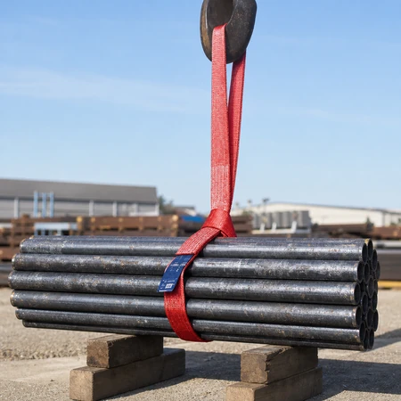 Red endless flat webbing sling hoisting a bundle of steel pipes from a storage yard with a crane